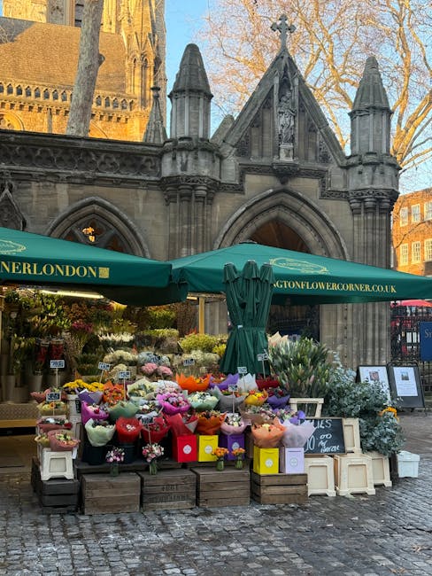 A vibrant outdoor flower stall situated in front of an historic stone church with Gothic architectural features, including pointed arches and pinnacles. The stall is covered by a large green canopy wi