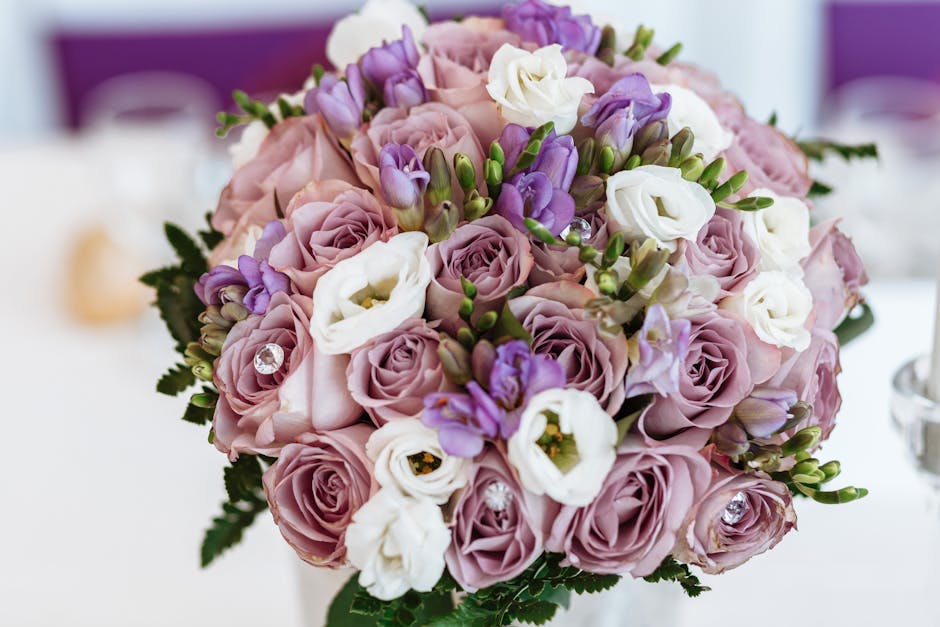 A close-up of a floral bouquet featuring soft pink roses, white lisianthus, and purple freesias arranged in a rounded bouquet style. The flowers are fresh with vibrant petals and green stems, adorned 