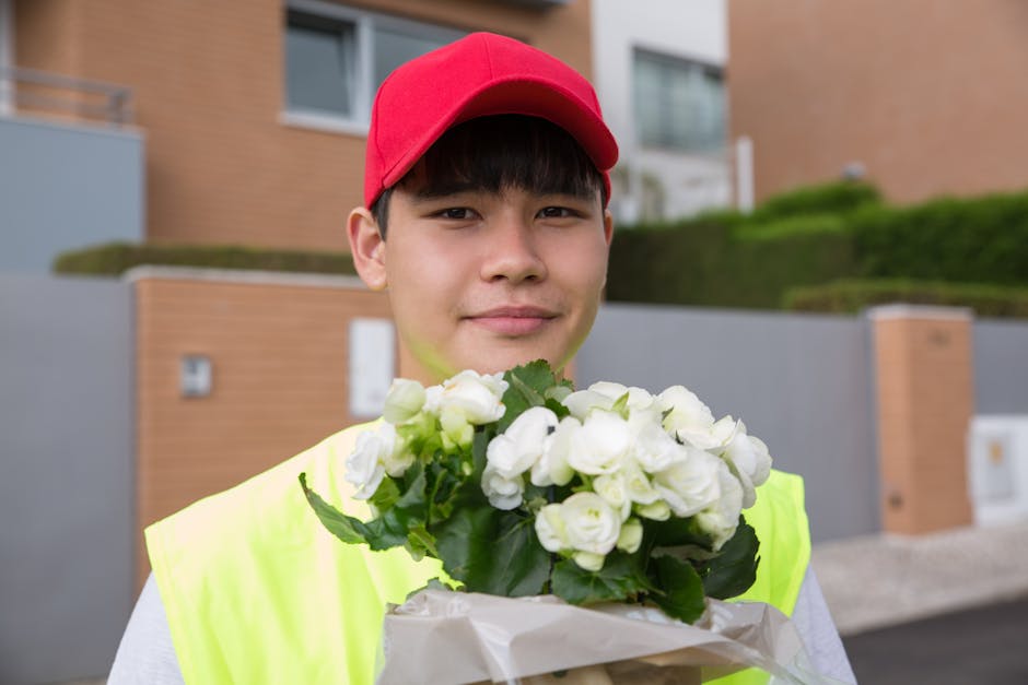 A young male flower delivery courier is standing outdoors in a modern residential area, holding a bouquet of fresh white roses with green leaves wrapped in white paper. He is wearing a red cap and a b