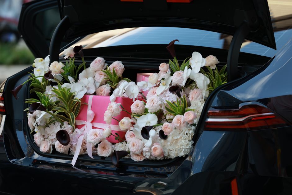 A car trunk filled with a lush floral arrangement composed of white orchids, pale pink roses, and dark purple calla lilies, complemented by green foliage. The flowers are arranged in a natural, slight
