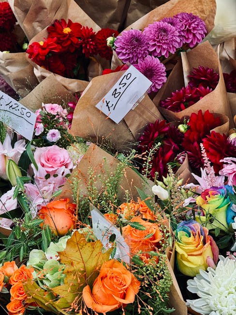 A vibrant assortment of fresh flowers arranged in small brown paper bouquets at a flower shop, with colors including bright red dahlias, pink and purple chrysanthemums, soft pink roses, orange roses,