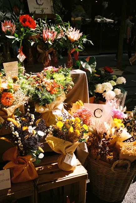 A display of vibrant fresh flowers arranged in various bouquets and containers at a flower shop, including pink lilies with delicate petals, bright red roses, orange gerbera daisies, and clusters of s