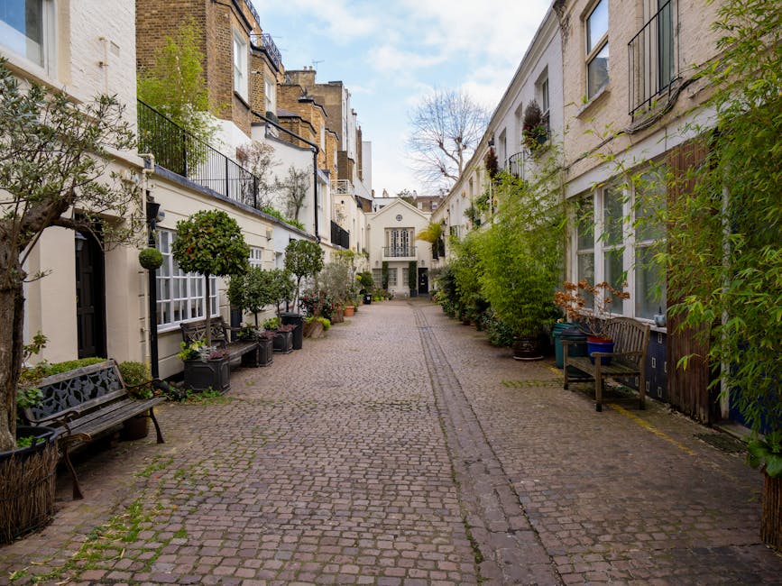 A narrow cobblestone alleyway flanked by white townhouse facades with black wrought-iron balconies and large windows. The scene features abundant greenery, including potted trees, shrubs, and climbing