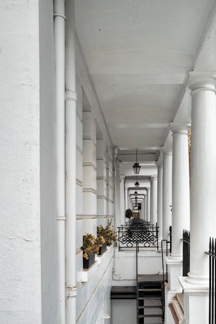This image shows a white architectural corridor featuring a series of evenly spaced, classical-style columns along the right side, supporting an overhead beam structure. The left side has a white wall