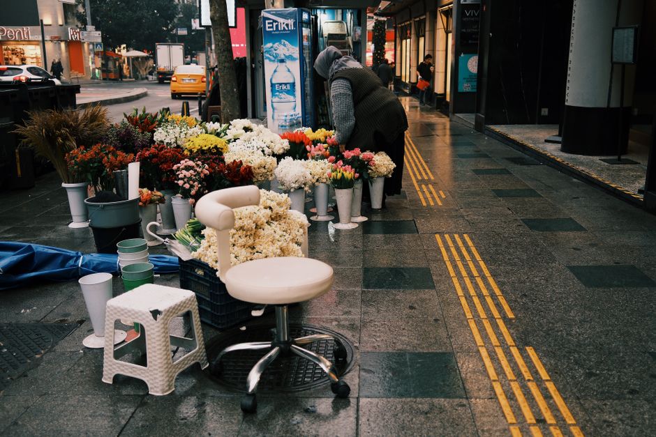 A street-side floral stall in SW7 displaying a vibrant assortment of fresh flowers, including white, pink, red, yellow, and orange blossoms arranged in white vases and buckets. The flowers include tul